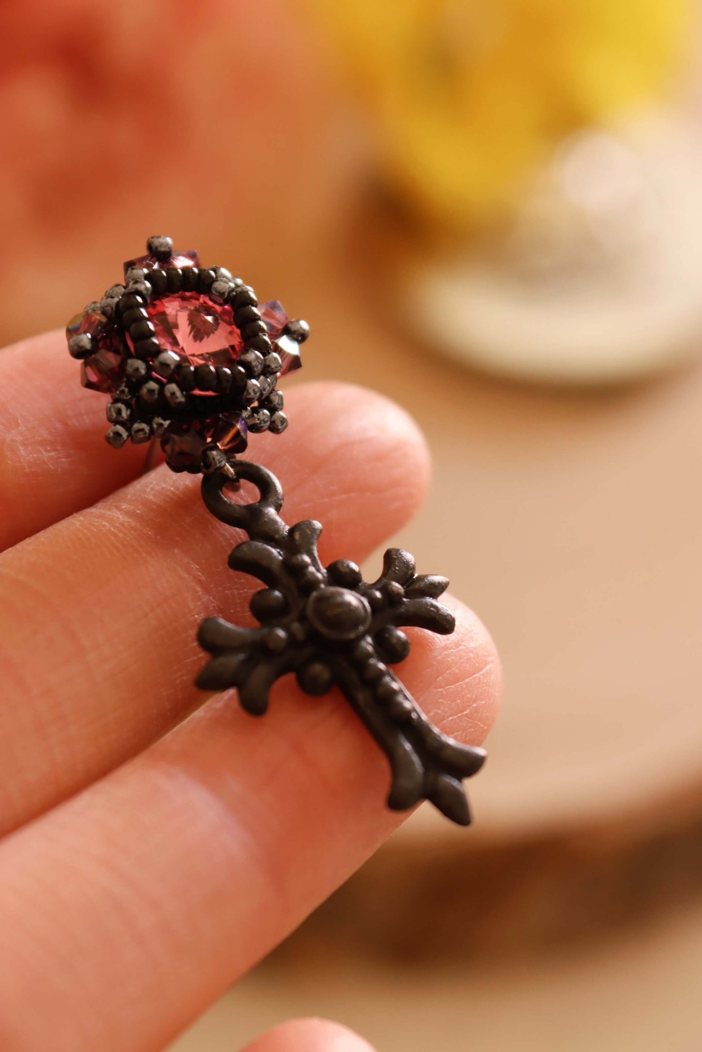 Close-up of a decorative black cross gothic earring and pink crystal held between fingers with a blurred background