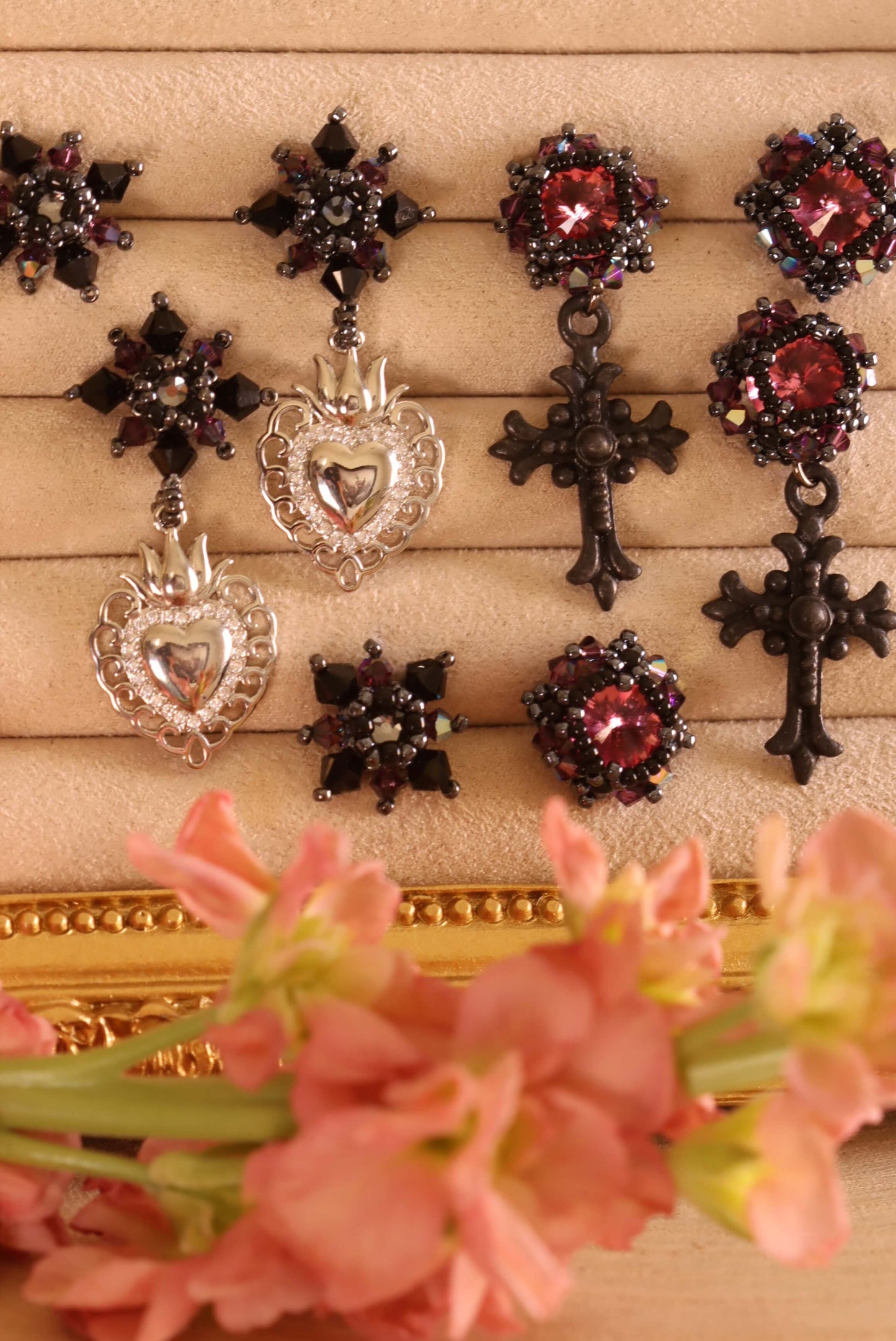 An overview of the Gothic Romance Jewelry Collection by Kaleidoscopes And Polka Dots - a collection of beaded earrings crafted with pink and black crystals, delicate beading, and stainless steel studs set against a beige background and flowers in the foreground.