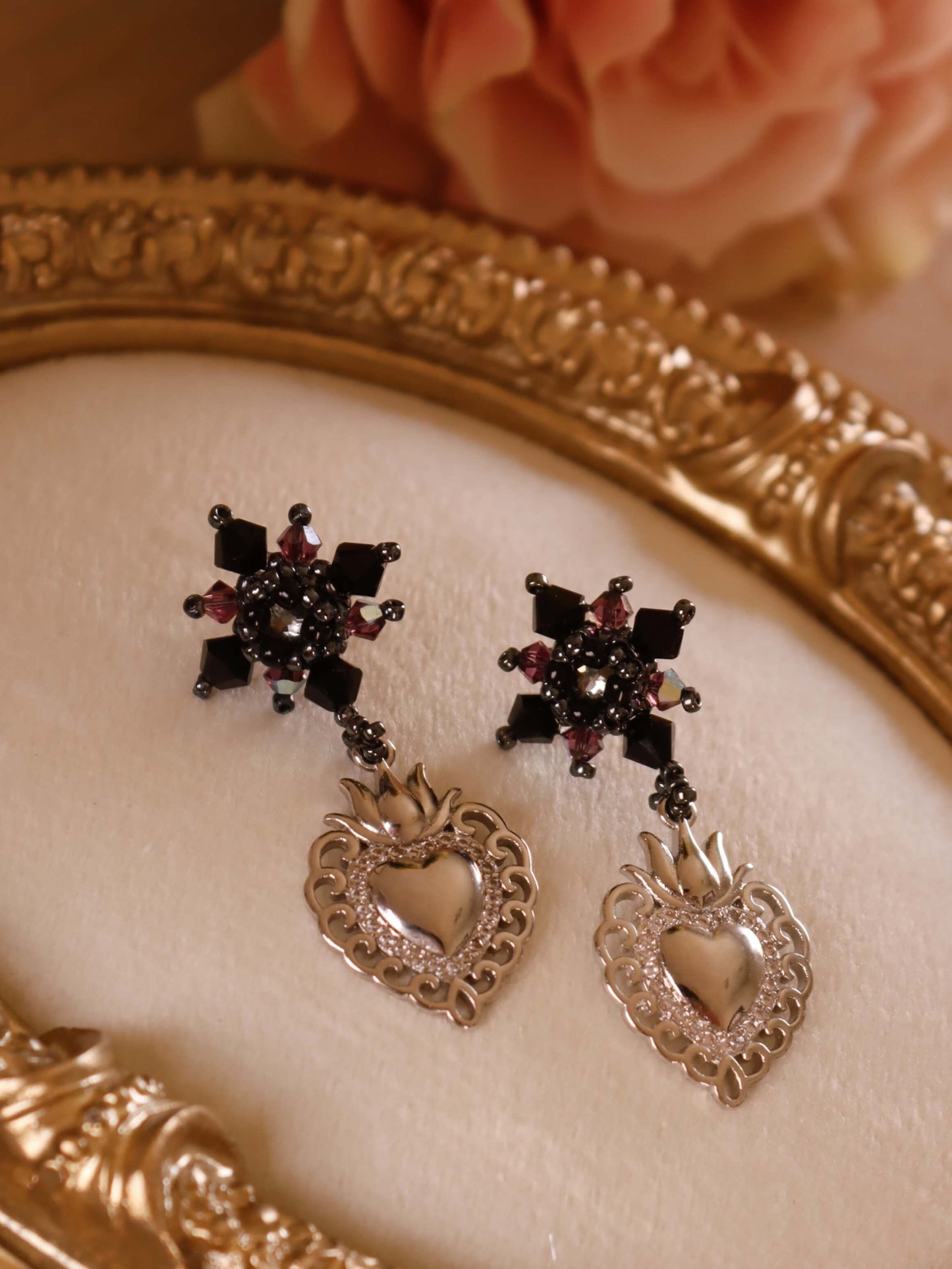 Sacred Heart Drop Earrings by Kaleidoscopes And Polka Dots are a pair of black crystal beaded earrings with elegant heart charms set against a decorative jewelry stand and pink rose in the background.