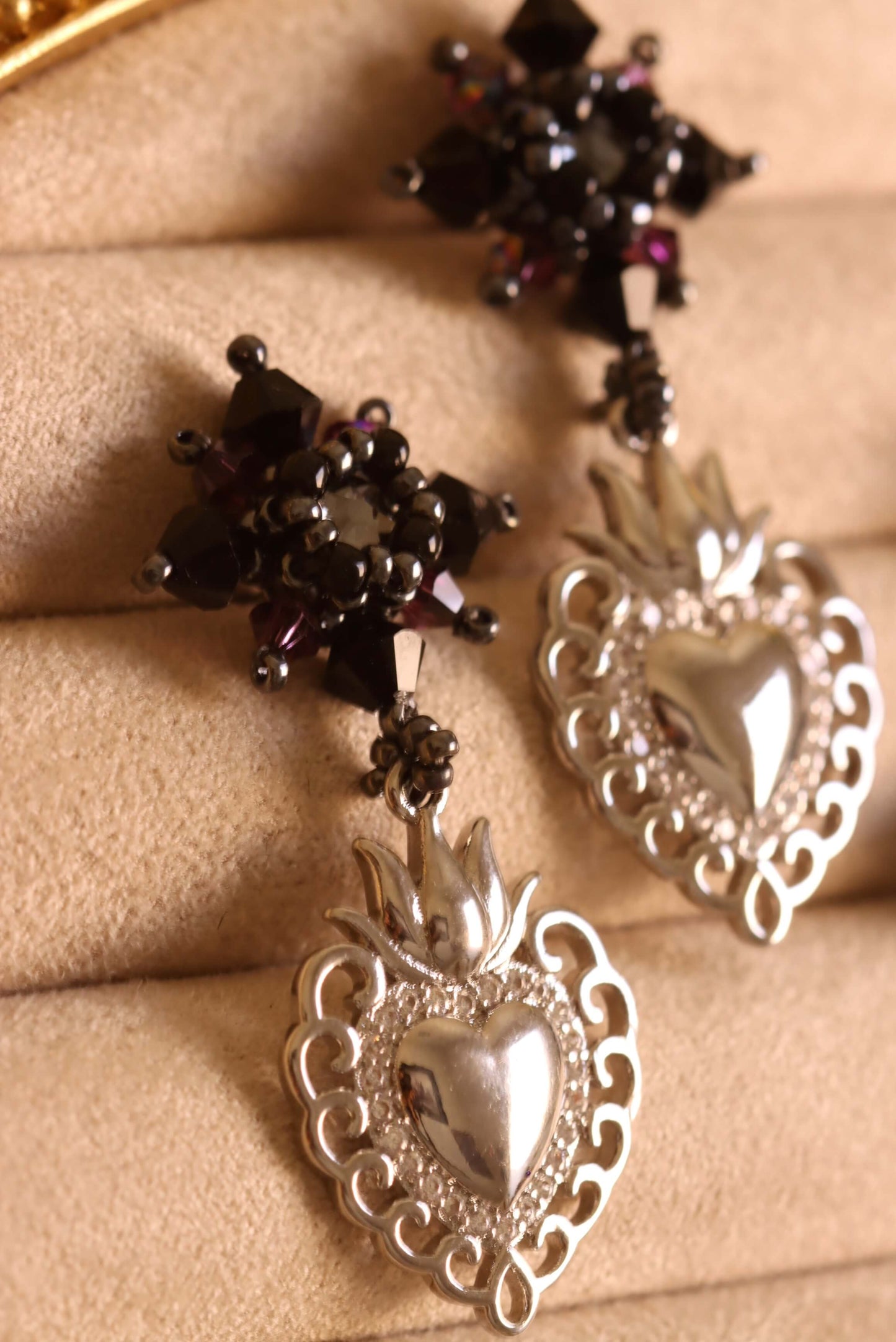 A detailed close-up shot of the Sacred Heart Drop Earrings by Kaleidoscopes And Polka Dots. This pair of elegant black crystal beaded earrings features silver heart charms set against a decorative jewelry stand.