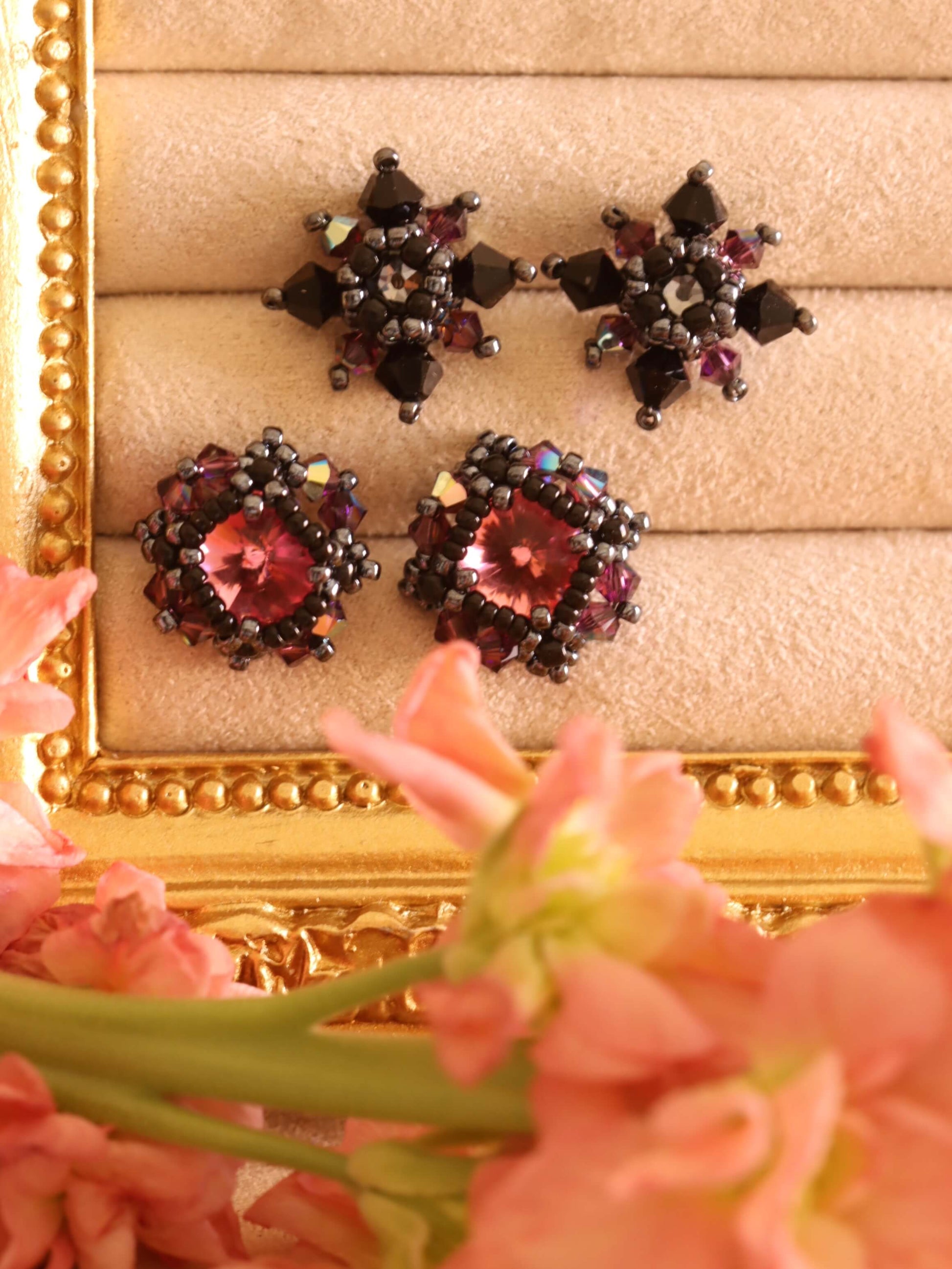 The Gothic Romance Jewelry Collection by Kaleidoscopes And Polka Dots features two sets of decorative stud earrings. These are displayed on a decorative stand with flowers in the foreground