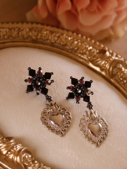 Sacred Heart Drop Earrings by Kaleidoscopes And Polka Dots are a pair of black crystal beaded earrings with elegant heart charms set against  a decorative jewelry stand and pink rose in the background.